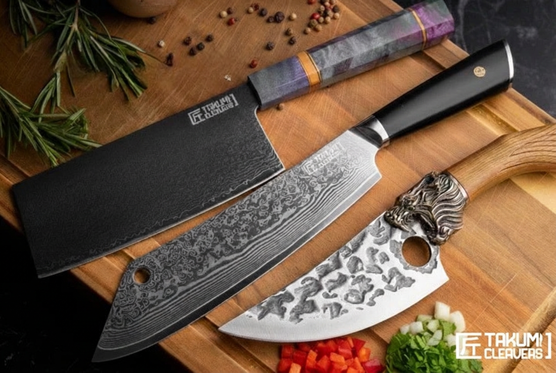 Three kitchen knives displayed on a wooden cutting board with herbs and chopped vegetables. The set includes a large cleaver-style knife with a black textured blade and black handle, a smaller santoku-style knife with a patterned silver blade and silver handle, and a utility knife with a purple marbled handle.