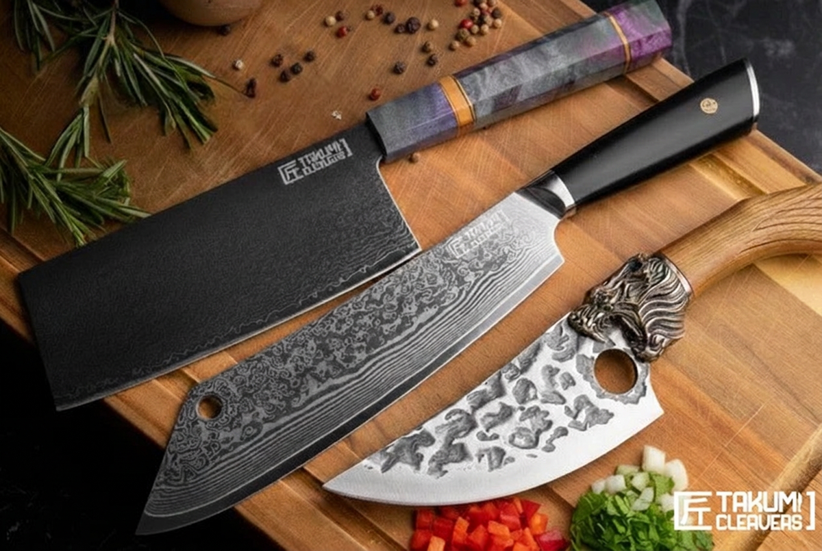 Three kitchen knives displayed on a wooden cutting board with herbs and chopped vegetables. The set includes a large cleaver-style knife with a black textured blade and black handle, a smaller santoku-style knife with a patterned silver blade and silver handle, and a utility knife with a purple marbled handle.